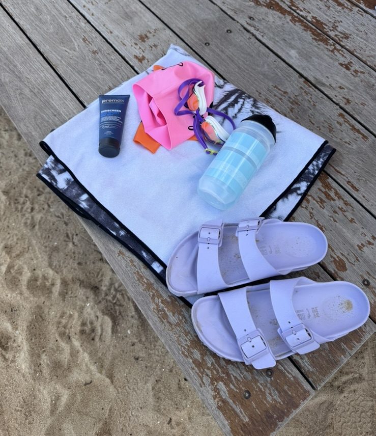 Swimming gear on a bench at the beach including towel, goggles, water bottle and sunscreen