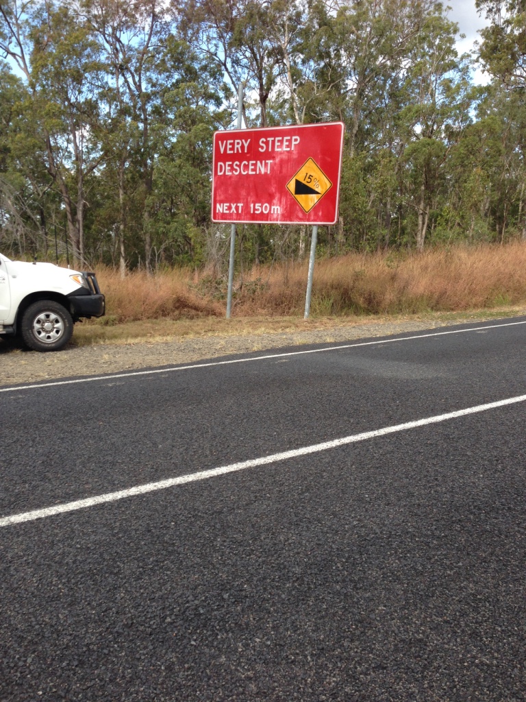 Roadside sign warning of a very steep descent ahead on a rural road.