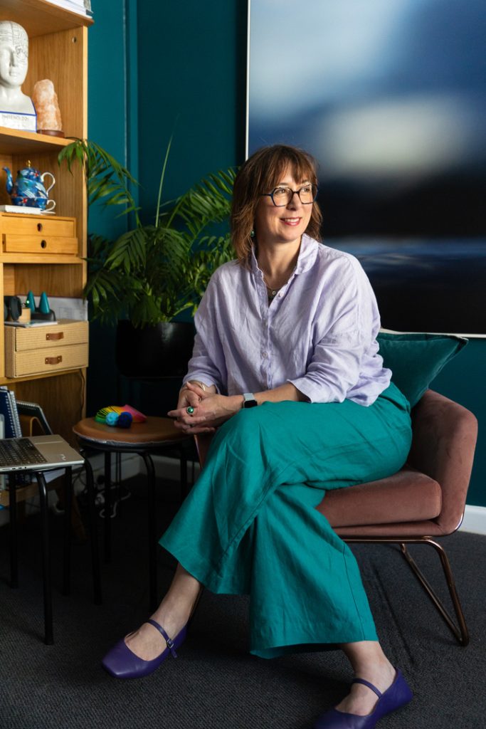 Nonie Carr seated in a therapy room, looking outward, representing reflective distance from self-critical thoughts