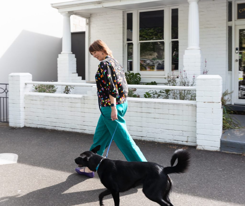 A woman walking a black dog along a sunny inner Melbourne street, relaxed and smiling