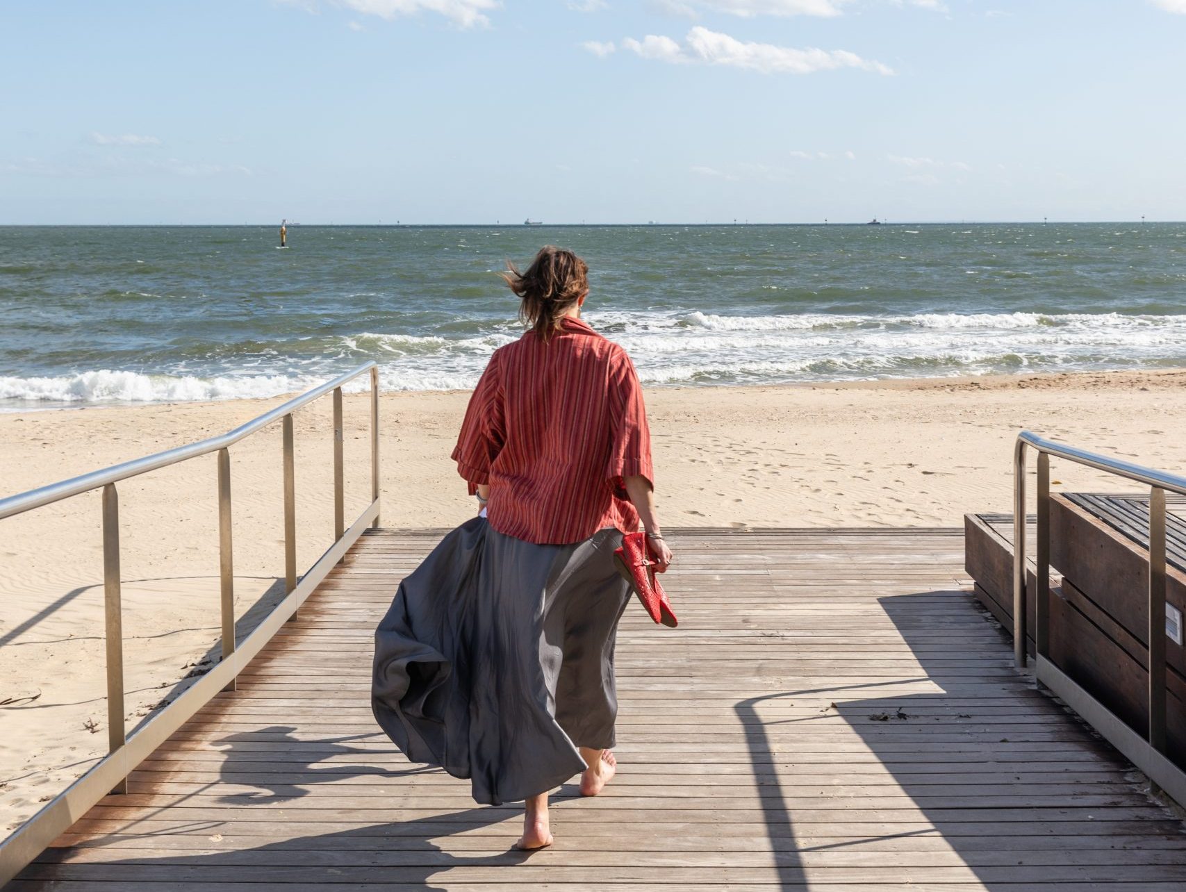A woman walking barefoot on a beachside boardwalk toward the open ocean, skirt moving in the wind