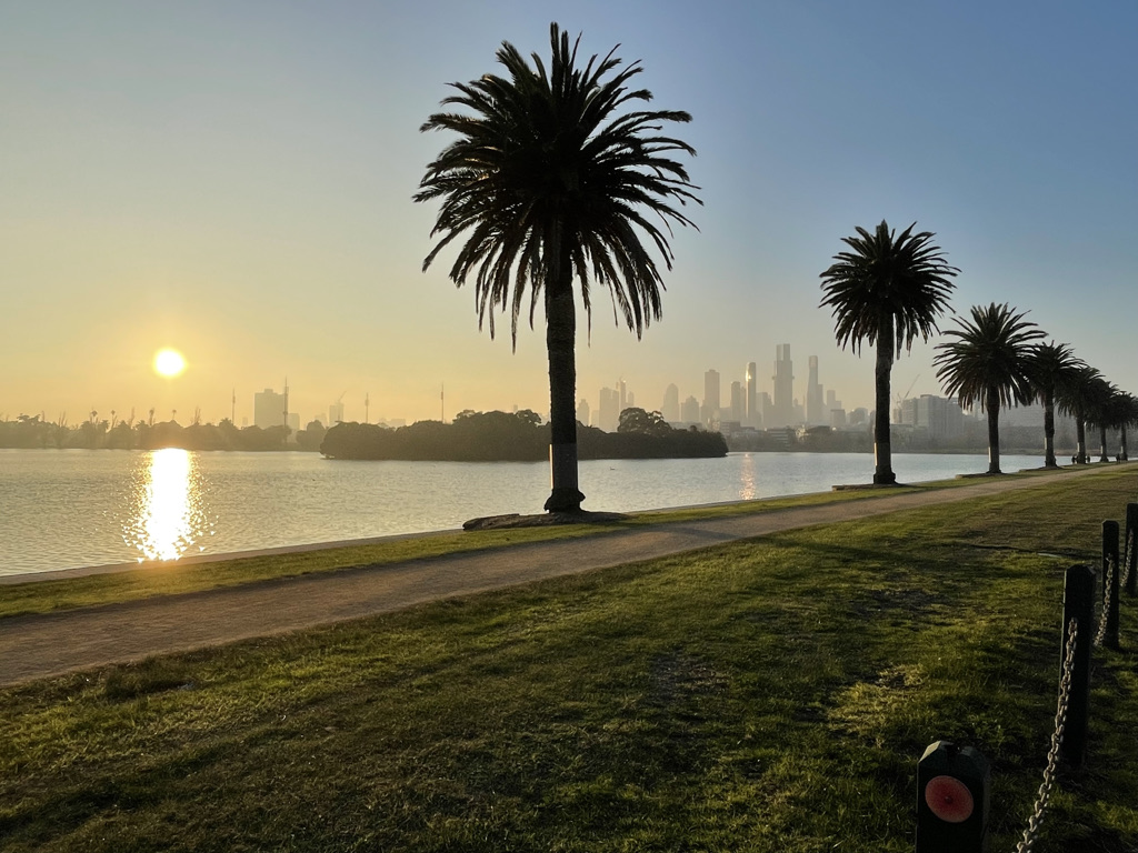 Sunrise over Albert Lake in Albert Park, Melbourne, palm trees silhouetted against golden morning haze, city skyline in the background, empty path along the water's edge