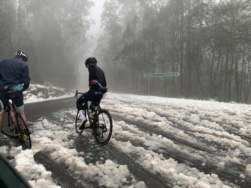 Two cyclists on a snow and slush covered road in foggy conditions near Warburton, Victoria, continuing to ride in difficult winter weather