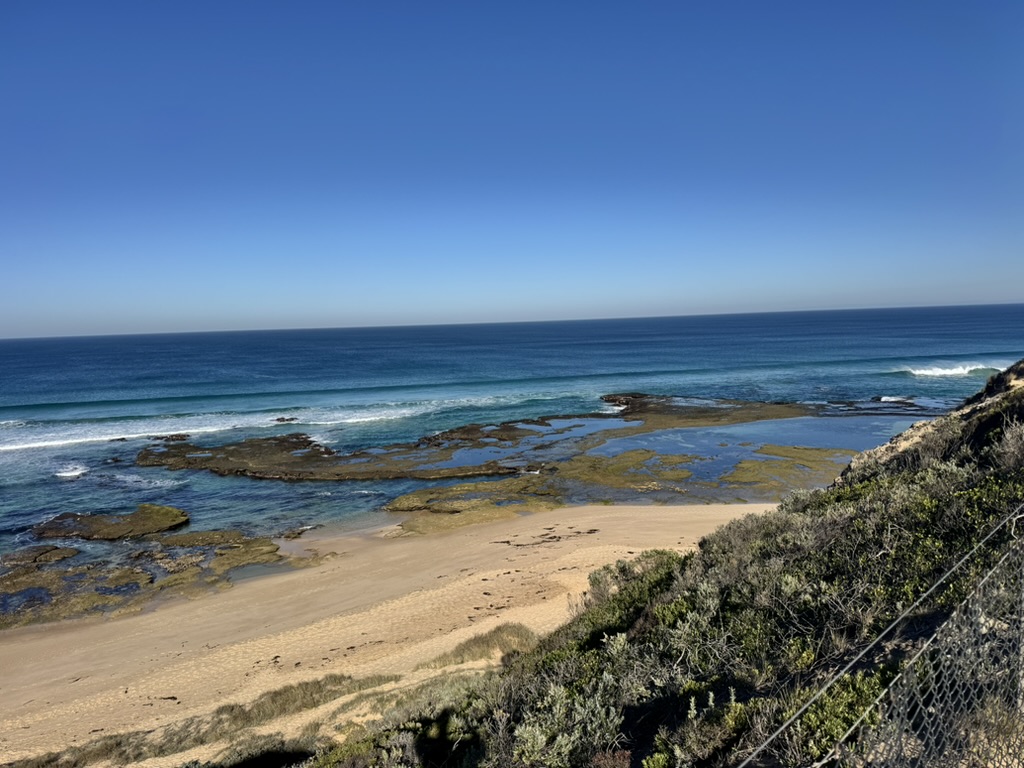 A wide, calm beach under an open blue sky with scattered cloud, the sea flat and still.