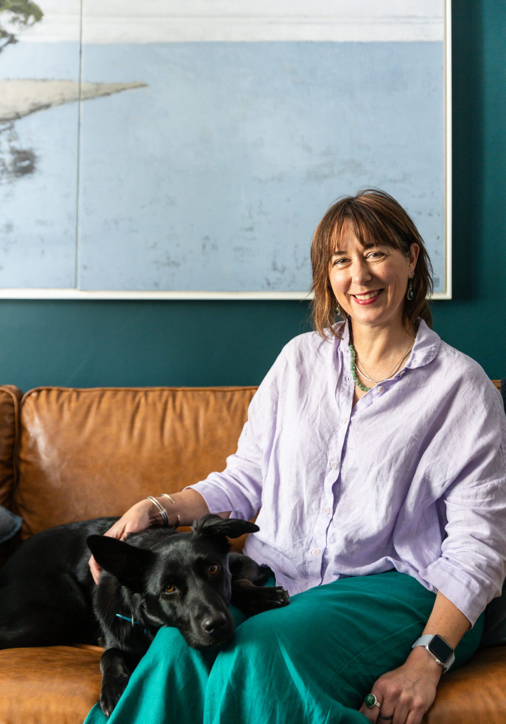Psychologist Nonie Carr sitting on a tan leather couch with her black kelpie, Bindi, at Enhance Life Psychology in Albert Park, reflecting a calm and welcoming therapeutic space.